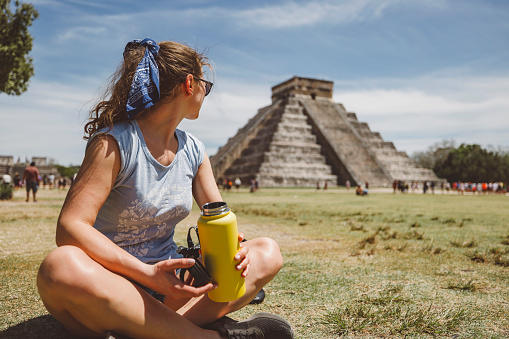 wWoman sitting on the ground holding a water bottle looking back at Chicken Itza.