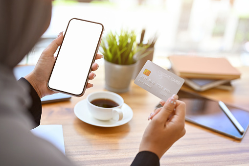 Close-up image, A muslim woman holding a smartphone and a credit card over the table. Phone white screen mockup. Online shopping, internet banking concept.