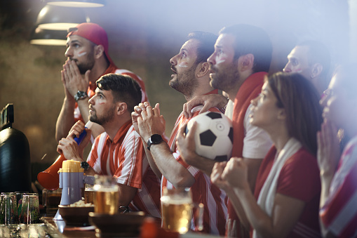 Large group of soccer fans watching the game of their team on TV in a bar.