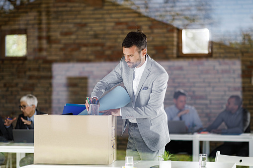 Mid adult businessman packing his belongings in the office after being fired from his job. The view is through glass.