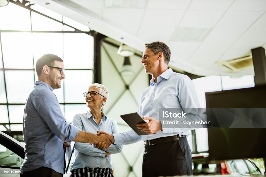 Business colleagues came to an agreement with car salesperson in a showroom. Happy businessman shaking hands with car salesperson while being in a showroom with his female colleague. Sales Occupation Stock Photo
