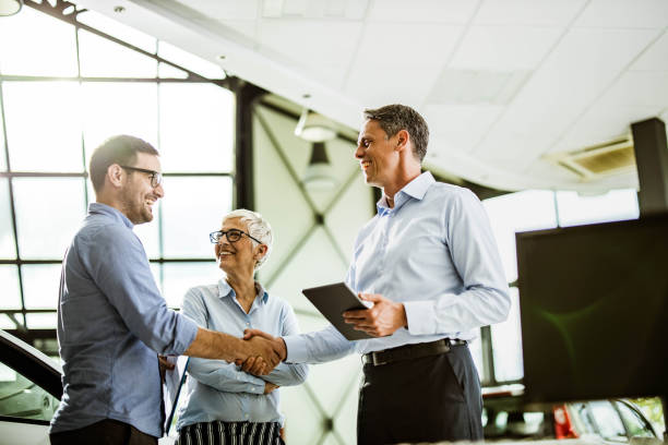 Business colleagues came to an agreement with car salesperson in a showroom. Happy businessman shaking hands with car salesperson while being in a showroom with his female colleague. salesman stock pictures, royalty-free photos & images