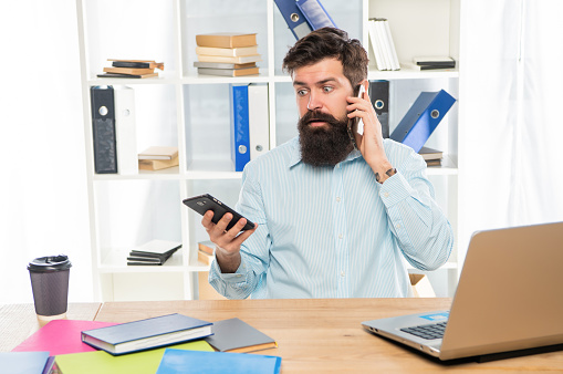 Shocked businessman talking on cellphone using smartphone at office desk, mobility.