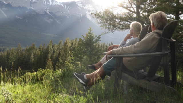 Couple relax on wooden chairs, look out over forest, mountains