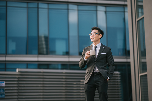 Portrait successful Asian Chinese Businessman holding coffee cup looking away outside office building