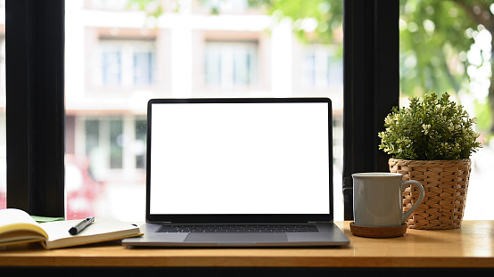 Vista frontal ordenador portátil, taza de café, planta de interior y cuaderno sobre mesa blanca. Lugar de trabajo cómodo. photo