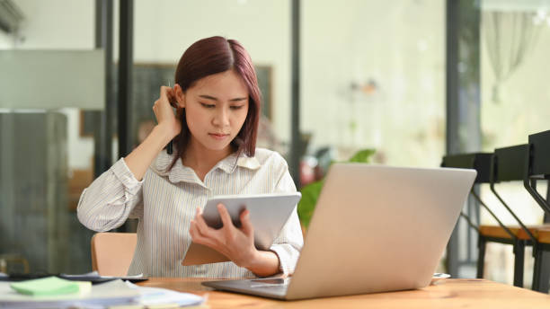 istock Focused asian female office worker sitting front of laptop computer and checking information on digital tablet. 1404818139