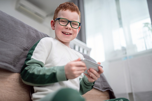 Portrait of cheerful boy with eyeglasses sitting on the floor and playing games on smartphone