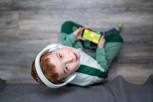 Cute joyful red-haired boy sitting on the floor at the bed and playing games on the smartphone