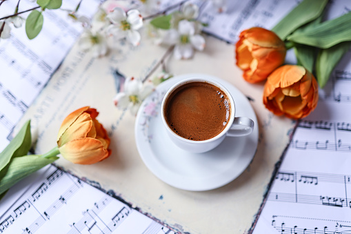 Istanbul, Turkey-June 20, 2022: Turkish coffee with plenty of foam in a white cup on mixed sheet music. Around the coffee are orange tulips and branches of white flowers. At the bottom of the coffee cup is an iron plate with a cream color vintage pattern. Shot with Canon EOS R5.
