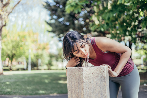 young indian woman drinking water in park fountain after jogging