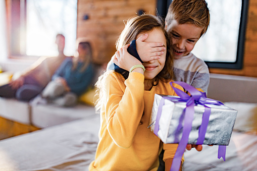 Happy little boy covering his sister's eyes while giving her a present at home. Their parents are in the background. Focus is on girl.