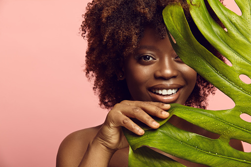 Cheerful young afro ethnicity woman and tropical leaves. Eco friendly cosmetics and beauty products for body and face care concept.