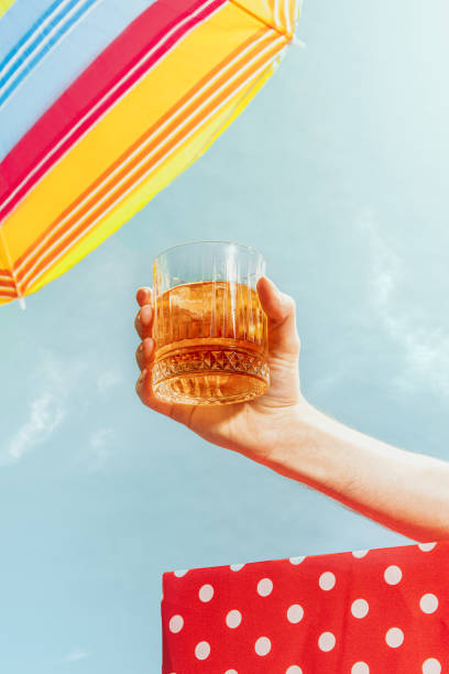 human hand holding glass of whiskey over summer blue sky background. vacation, happiness, summer vibes and ad concept. retro style - celebration imagens e fotografias de stock