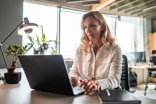 Attractive mature caucasian businesswoman and entrepreneur sitting and working in the office. She is wearing casual clothes and using a laptop.