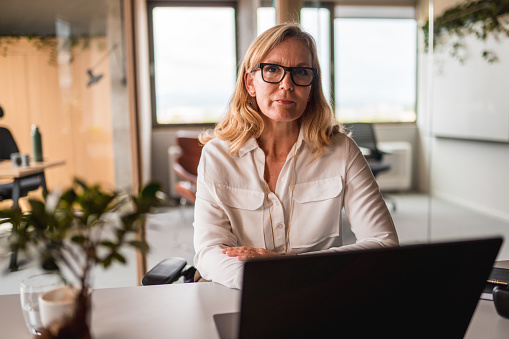 Waist up front view of a beautiful mature caucasian female businesswoman, sitting at her desk in front of a laptop. She is wearing eyeglasses and looking at the camera.