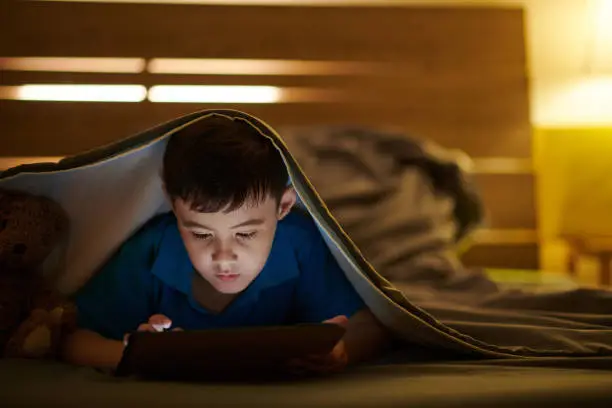 Preteen boy lying under blanket and playing game on tablet computer late at night