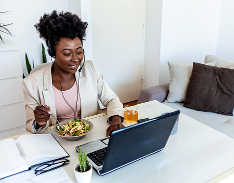 Beautiful afro american young woman sitting at the desk in an office and eating lunch, using laptop at the same time. Close up of a young woman having lunch in her home office.