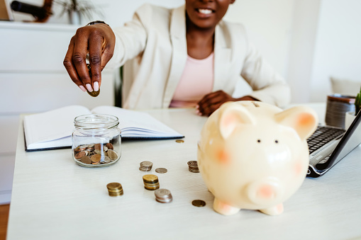 Woman hand putting money coin into piggy for saving money wealth and financial concept. Closeup shot of an unrecognisable businesswoman filling a glass jar with coins in an office.