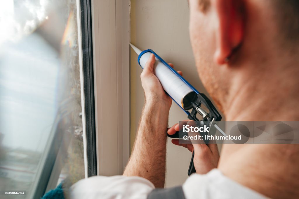 Young man wearing overalls sealing cracks between window and trim Young man wearing overalls sealing cracks between window and trim using waterproof silicone caulk on the balcony. Window Stock Photo