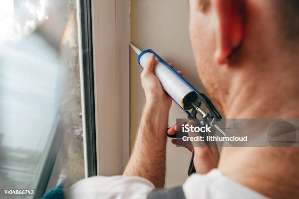 Young Man Wearing Overalls Sealing Cracks Between Window And Trim Stock Photo - Download Image Now