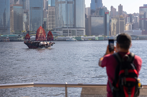 Hong Kong - June 23, 2022 : A man takes a photo of a junk boat with a slogan celebrating the 25th anniversary of Hong Kong's handover from Britain to China, on Victoria Harbour, Hong Kong.