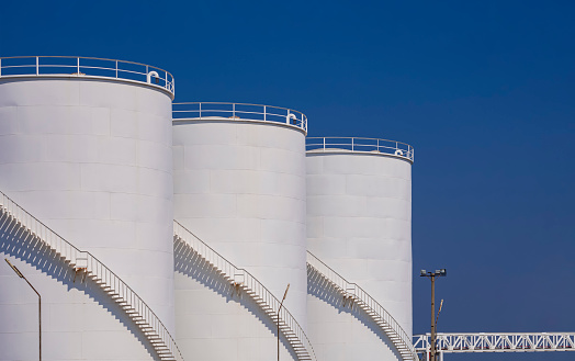 Three white storage fuel tanks with oil pipeline system in petroleum industrial area at harbor against blue clear sky background