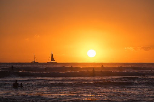 Oahu, USA - Nov 6, 2021: Sunset off Waikiki beach with sail boats and surfers catching waves.
