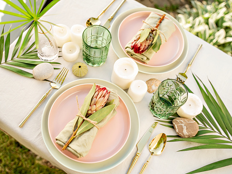 Festive table setting for two person in tropical style. Restaurant, wedding, party decorated with flowers, candles and leaves. Midsummer mood event, romantic dinner. Florist decorator occupation.