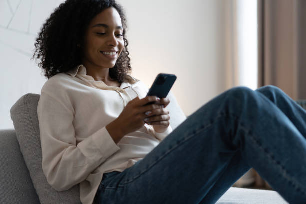 istock Smiling young mixed race woman sit on sofa at home using cellphone texting chatting with friend 1404882180