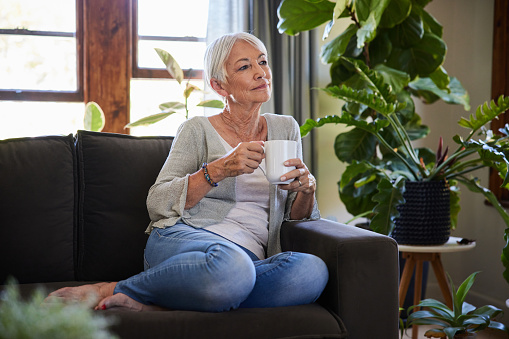 Senior woman drinking a cup of coffee and looking lost in thought while sitting on a sofa in her living room at home