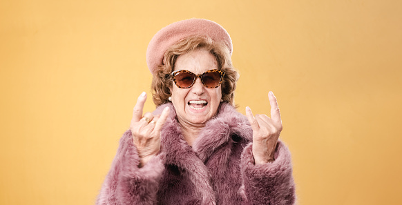 Happy senior female in trendy pink fur coat and hat with sunglasses gesturing horns and smiling against yellow background
