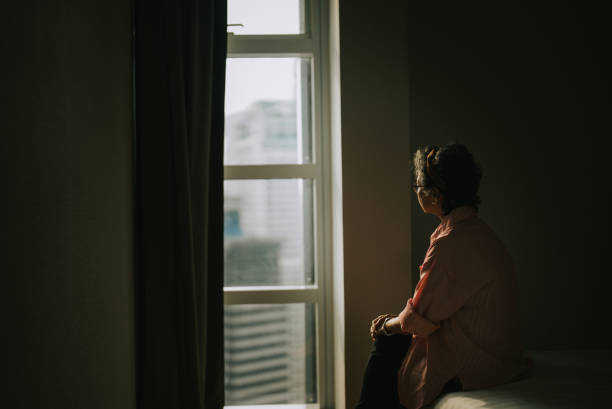 lonely asian chinese senior woman standing in the dark behind the window looking stock photo