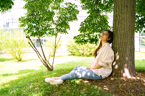Young Asian Thai, Vietnamese or Chinese woman in casual clothes and glasses sitting relaxing and dreaming in shadow under green tree on weekend, holiday or vacation in city park on summer sunny day.