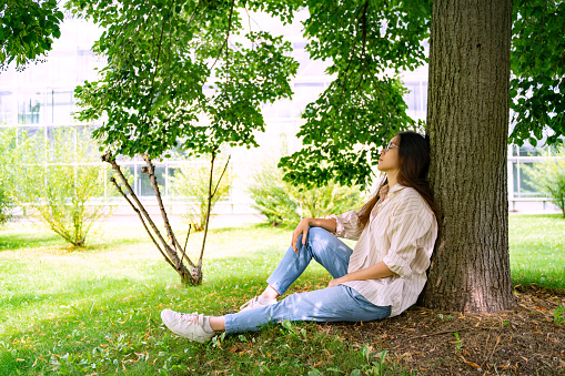 Young Asian Thai, Vietnamese or Chinese woman in oversize shirt, jeans and glasses sitting relaxing recreating in shadow under green tree on weekend or holiday in city park on summer sunny day.