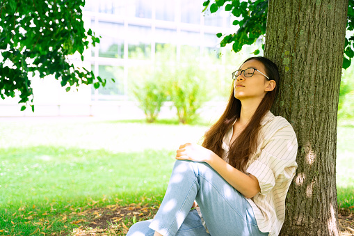Young Asian Thai, Vietnamese or Chinese woman in casual clothes and glasses sitting with closed eyes relaxing and sleeping in shadow under green tree on weekend in city park on summer sunny day.