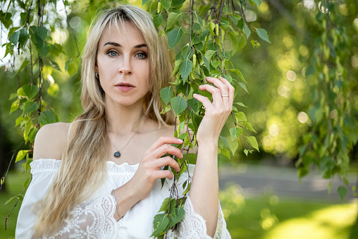 Young blonde woman wearing white dress posing among birch tree branches with green leaves