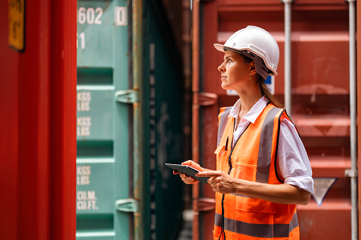 Female manager wearing reflective vest and hard hat using tablet working at container yard. Container yard warehouse worker. Cargo Shipping Import and Export industry. Logistic shipping yard business.