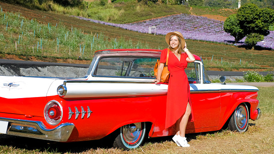 Happy woman in red dress enjoy sunset near country road and classic Vintage sports car. Romantic freedom travel concept. Road trip