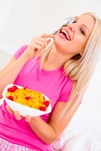 Silly Smiling Caucasian woman in pink pajamas eating cornflakes cereal sitting in bed.
