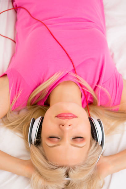 Above view of Smiling Caucasian woman in pink pajamas listening to headphone laying in bed.