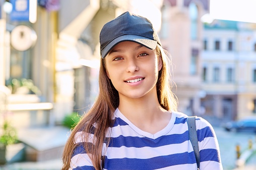 Outdoor portrait of teenage girl in black cap, city street background. Beautiful fashionable confident female looking at camera, in sunset light. Adolescence, style, lifestyle, beauty, youth concept