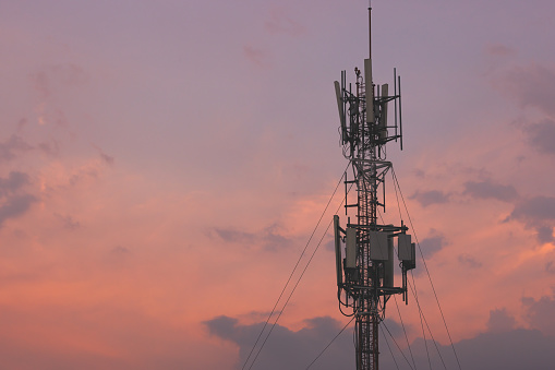 Telecommunication tower and its wireless cellular transmission antenna in orange sunset sky background. Mobile phone data network transmitter technology and cell site concept.