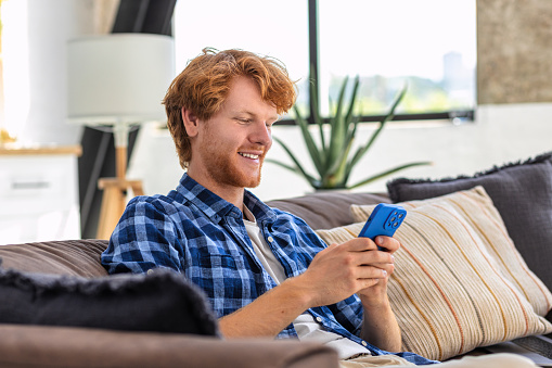 Young joyful man using mobile phone and smiling sitting at home on the couch. Redhead Male holds smartphone in his hands looking at the screen, reads a message, plays games