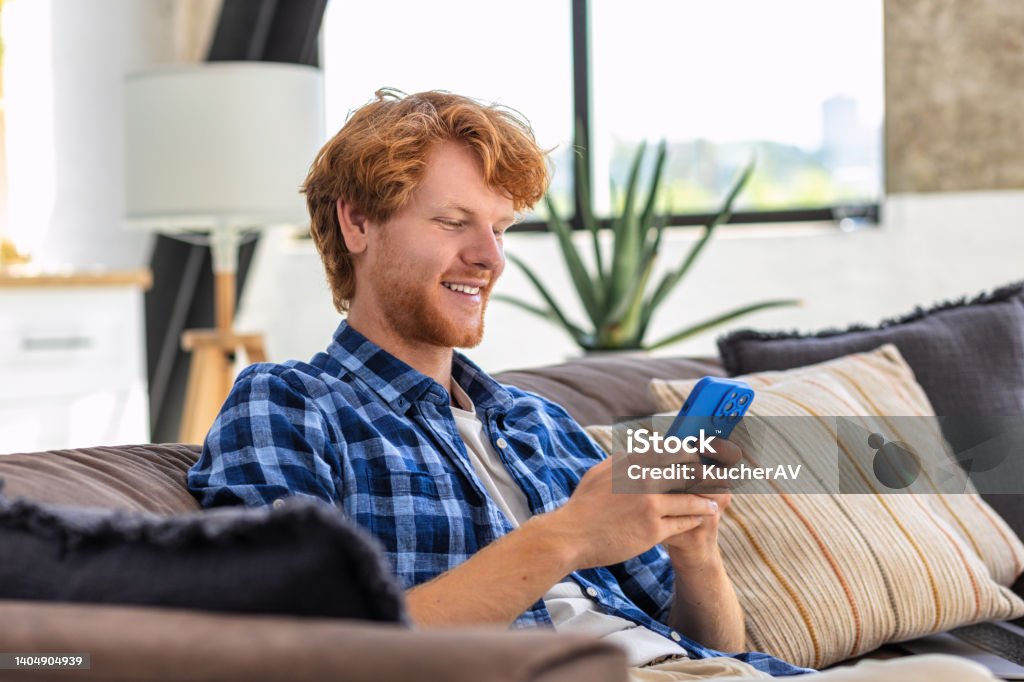 Young joyful man using mobile phone and smiling sitting at home on the couch. Redhead Male holds smartphone in his hands looking at the screen, reads a message, plays games Text Messaging Stock Photo