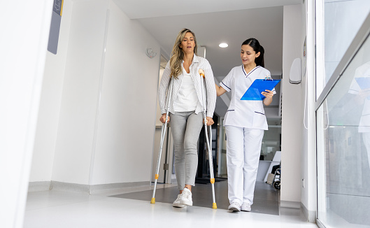Latin American nurse helping a female patient walking in crutches at the hospital - healthcare and medicine concepts