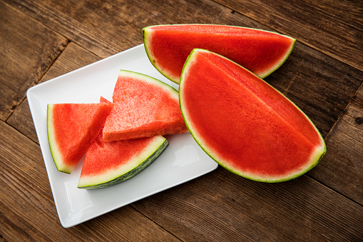 This is a high angle photograph of a plate of triangle shaped sliced cut watermelon on a white modern plate. It is surrounded by larger pieces sitting on a wood Beach deck