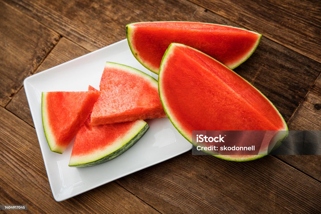 Triangle shaped sliced cut watermelon on a white plate This is a high angle photograph of a plate of triangle shaped sliced cut watermelon on a white modern plate. It is surrounded by larger pieces sitting on a wood Beach deck Watermelon Stock Photo