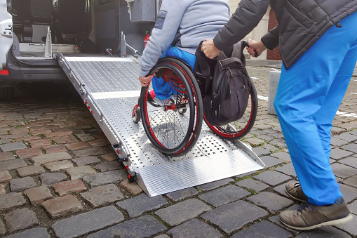 Person on wheelchair with disability using accessible car ramp for transport with help of an assistant driver.