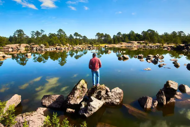 Photo of Man standing on a rock in front of a reflecting lake with a forest in the background on a sunny day in Mexiquillo natural park in Durango